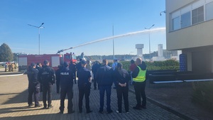 uczestnicy ćwiczenia stoja na placy i obserwują działania strażaków, którzy leją wodę na miejsce zagrozone